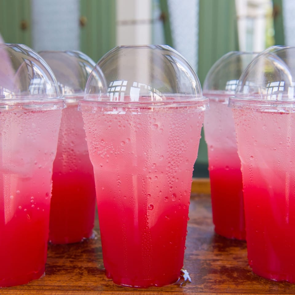 Several clear plastic cups filled with vibrant pink beverages, ice, and condensation on a wooden surface.