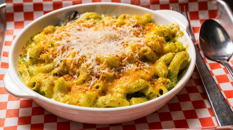 Baked pasta with green sauce, melted cheese, and breadcrumbs in a white dish on a red checkered paper.