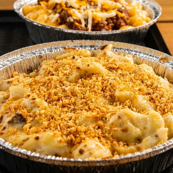 Two foil containers of mac and cheese, one topped with breadcrumbs, the other with chili and cheese.
