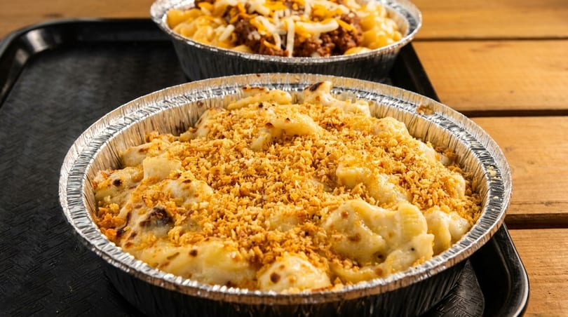 Two foil containers of mac and cheese, one topped with breadcrumbs, the other with chili and cheese.