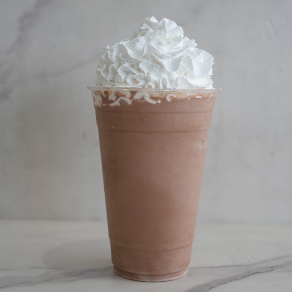 A chocolate milkshake in a clear plastic cup, topped with a generous swirl of whipped cream.