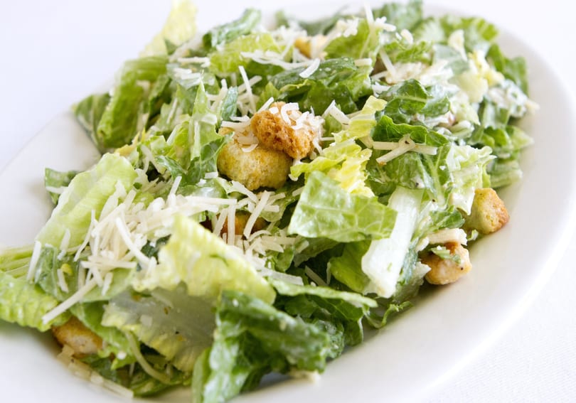 Close-up of a Caesar salad with romaine lettuce, croutons, and shredded Parmesan cheese on a white plate.