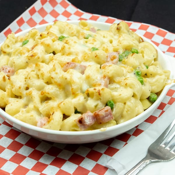 Baked macaroni and cheese with ham and peas in a white dish on a red and white checkered paper.