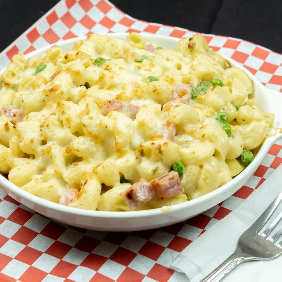Baked macaroni and cheese with ham and peas in a white dish on a red and white checkered paper.
