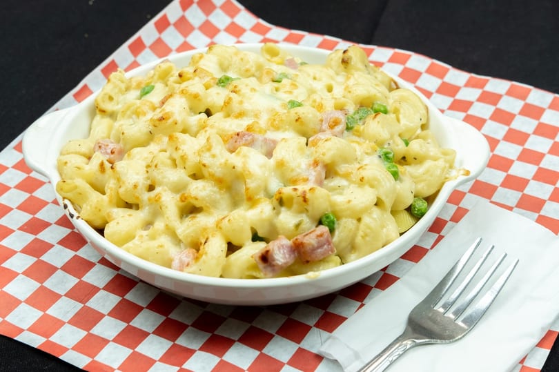 Baked macaroni and cheese with ham and peas in a white dish on a red and white checkered paper.