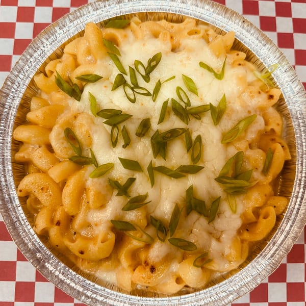 Macaroni and cheese in a foil tin, topped with melted cheese and green onions, on a red and white checkered background.