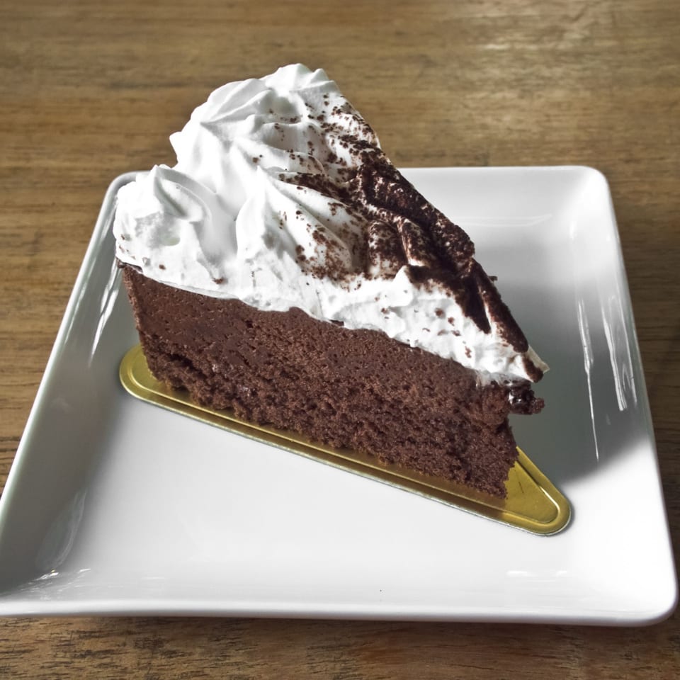 A slice of rich chocolate cake topped with whipped cream and cocoa powder on a white plate.