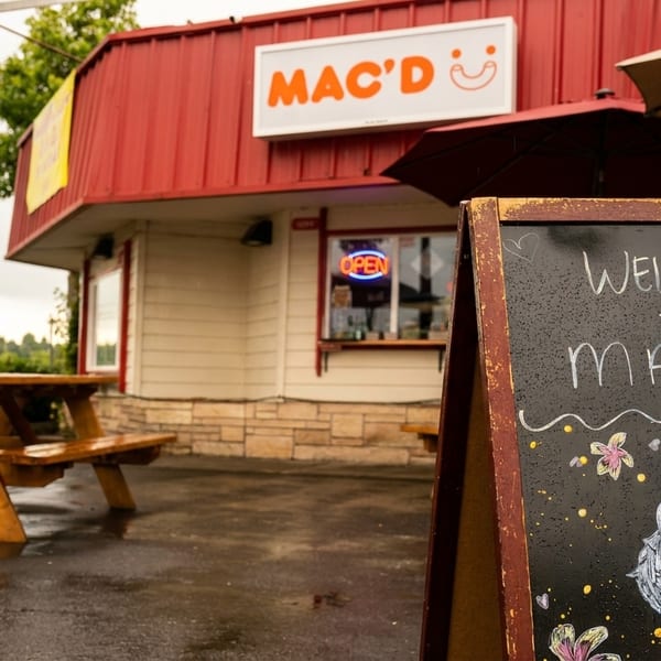 Mac'D restaurant exterior with a "Welcome to Mac'D" chalkboard sign and outdoor picnic tables.