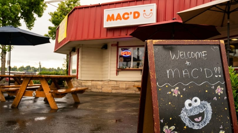 Mac'D restaurant exterior with a "Welcome to Mac'D" chalkboard sign and outdoor picnic tables.