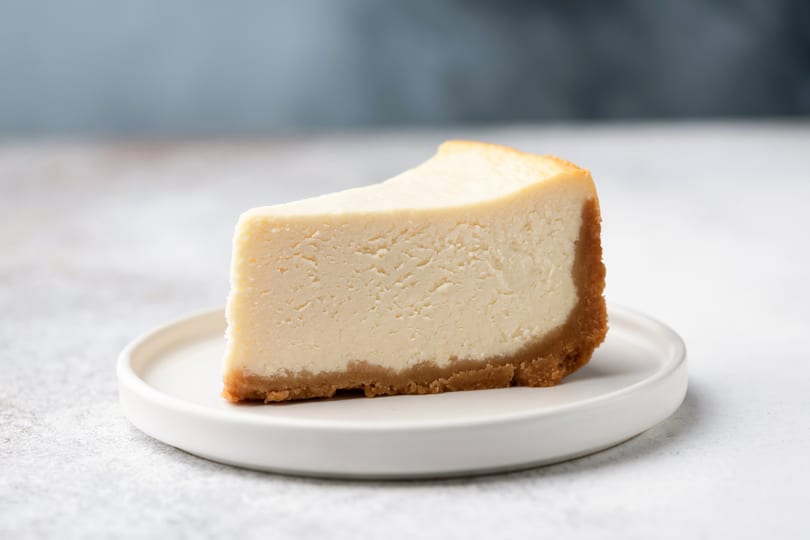 A slice of classic cheesecake with a graham cracker crust sits on a white plate.