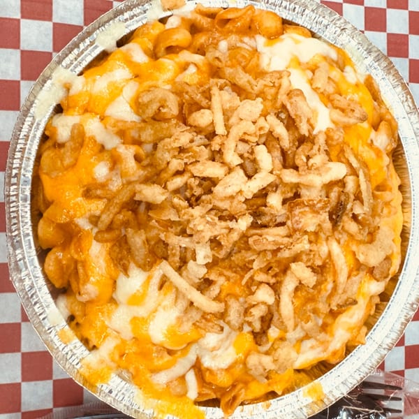 Macaroni and cheese topped with crispy fried onions in a foil container on a red and white checkered tablecloth.