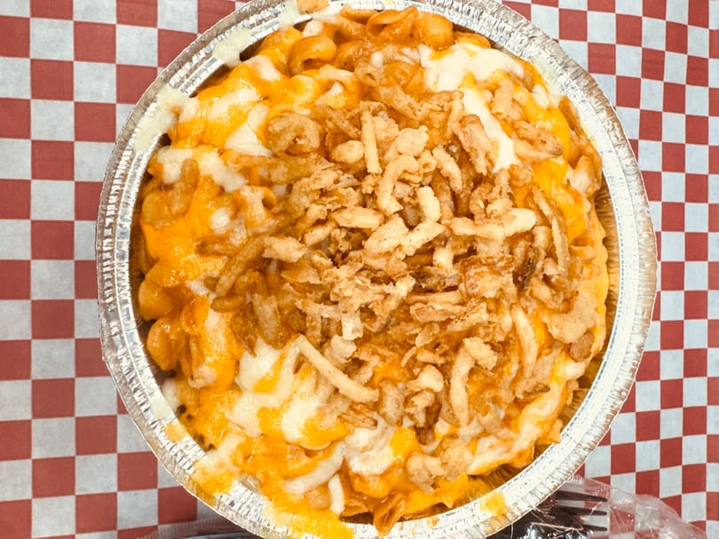 Macaroni and cheese topped with crispy fried onions in a foil container on a red and white checkered tablecloth.