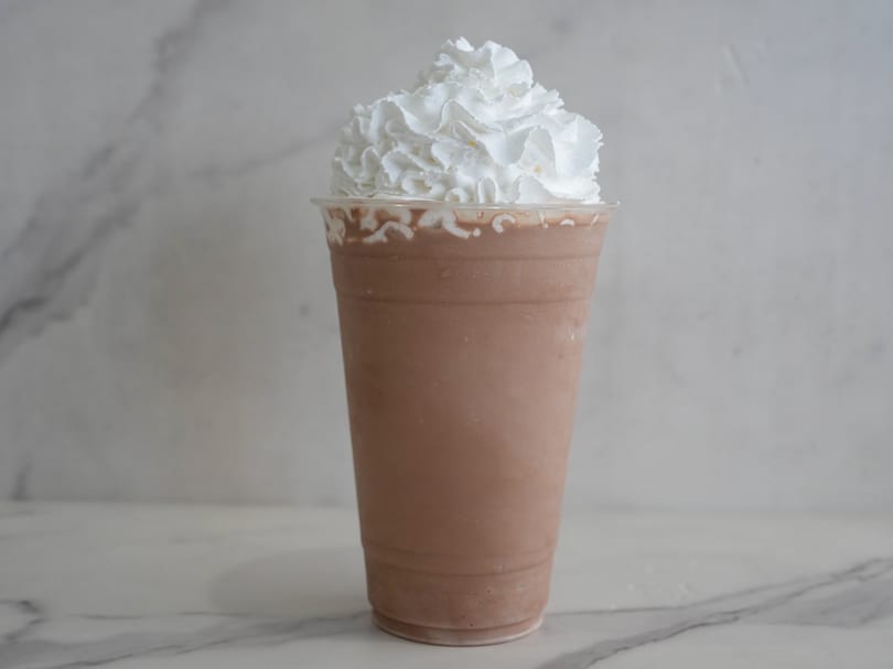 A chocolate milkshake in a clear plastic cup, topped with a generous swirl of white whipped cream.