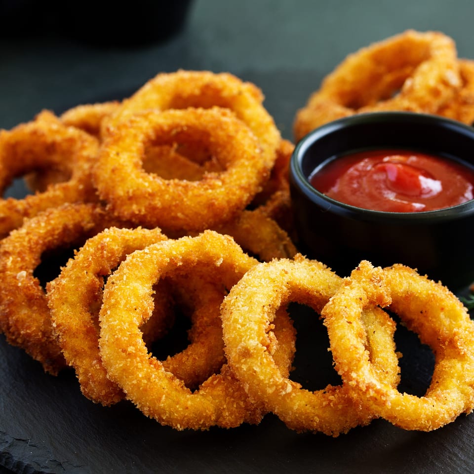 Golden-brown crispy onion rings piled on a dark slate board with a small bowl of ketchup.