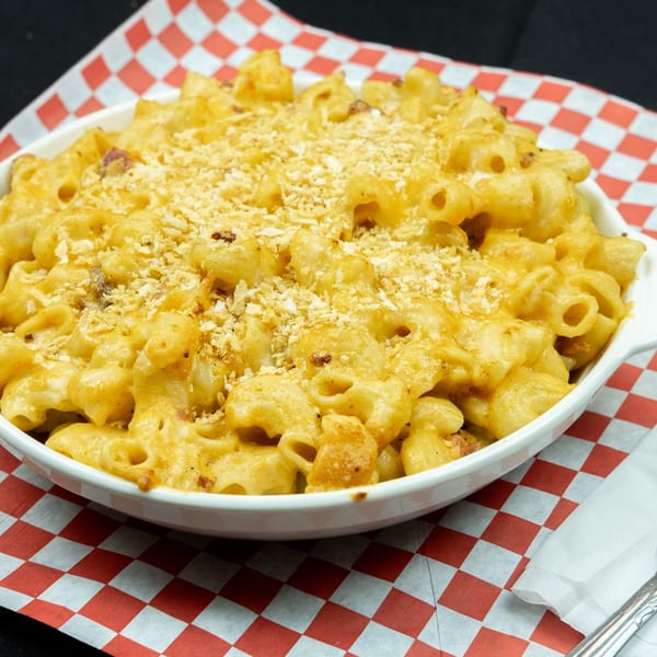 A white bowl of macaroni and cheese with breadcrumbs, on red and white checkered paper.