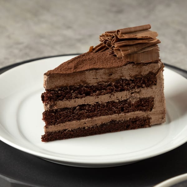 A slice of rich chocolate cake with chocolate frosting, cocoa powder, and chocolate shavings on a white plate.