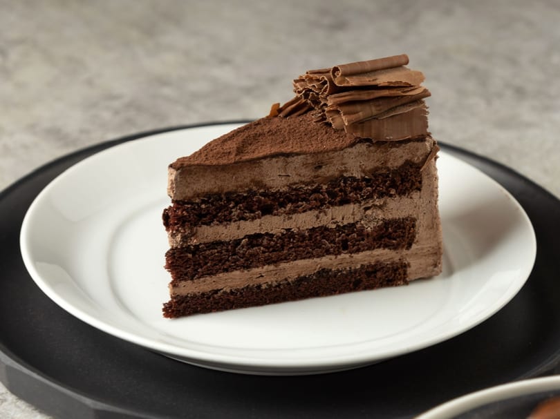 A slice of rich chocolate cake with chocolate frosting, cocoa powder, and chocolate shavings on a white plate.