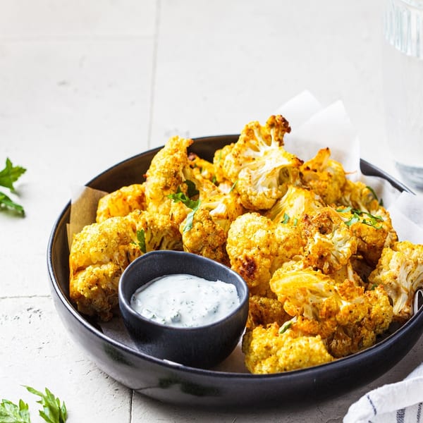Golden roasted cauliflower florets with a side of creamy herb dip in a black bowl.
